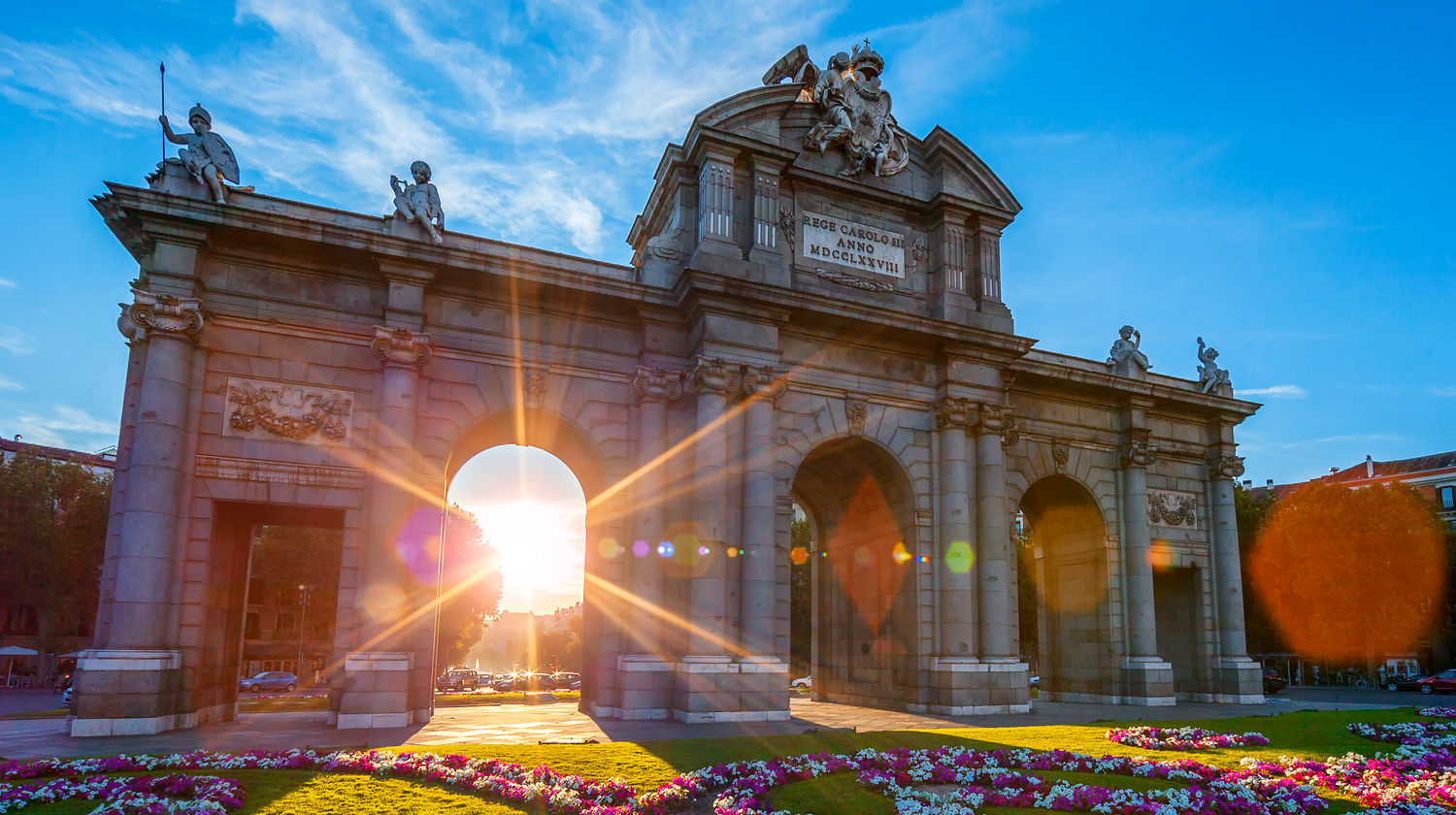 Vista da Puerta de Alcalá em Madrid ao nascer do sol, com a luz dourada a atravessar o arco e jardins floridos em primeiro plano