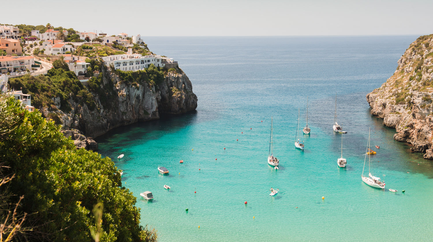 Aerial view of Cala Mitjana Beach in Menorca, Spain, showing turquoise waters, cliffs, and anchored boats along the Balearic coastline.