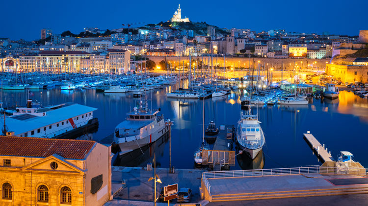 Night view of Marseille Old Port showing illuminated waterfront buildings, docked boats, and the city rising above the harbor in southern France