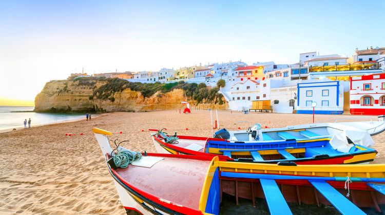 A sunset view of Carvoeiro in the Algarve, showing whitewashed houses perched on cliffs above a sandy beach with warm golden light reflecting on the ocean.