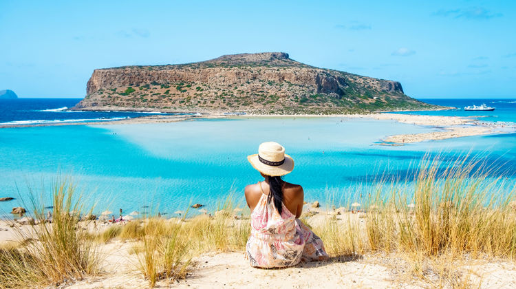 View of Balos Lagoon in Crete, Greece, with turquoise waters, sandy shoreline, and the rocky peninsula in the background.