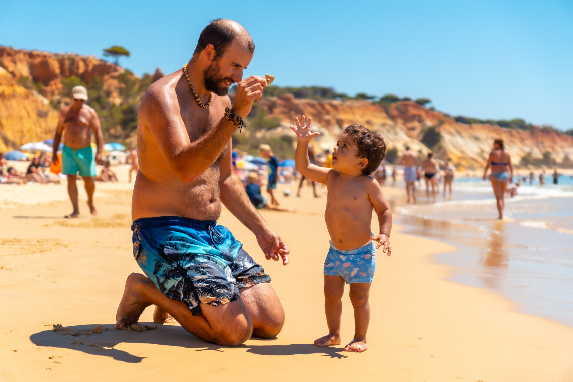 Father and child playing together on the golden sandy beach of the Algarve, Portugal, with cliffs and families in the background on a sunny summer day.