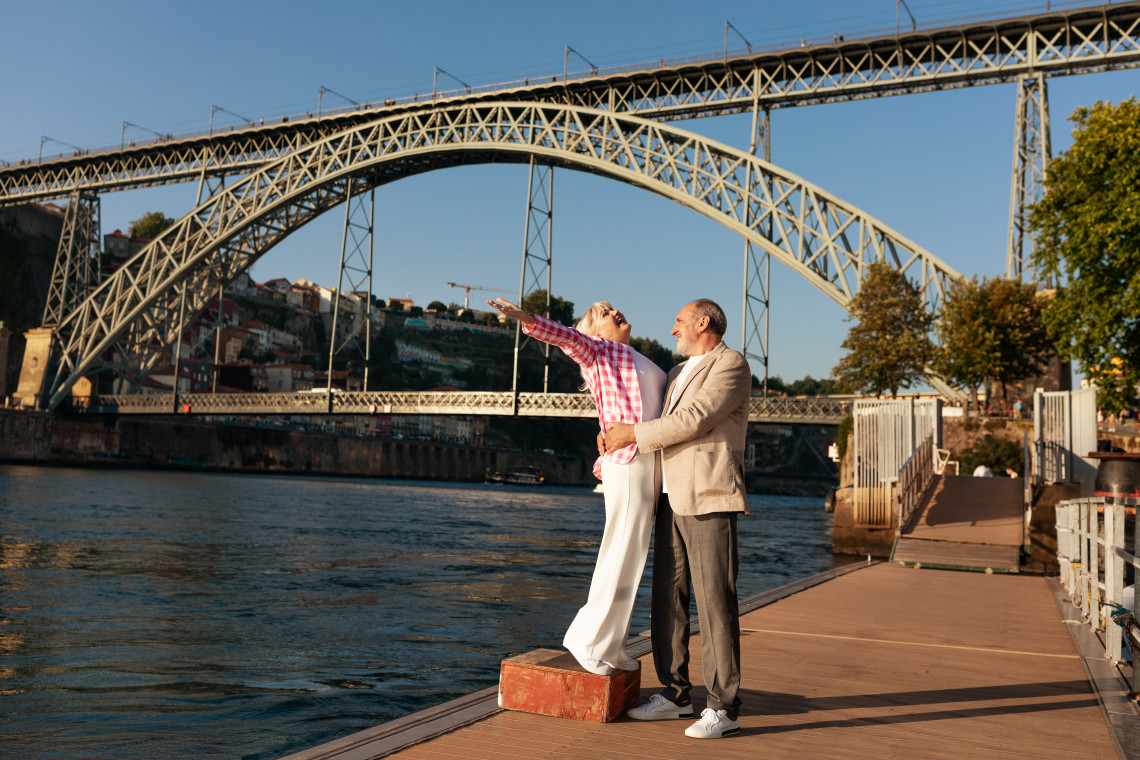 Happy couple standing by the Douro River with the Dom Luís I Bridge in the background at sunset, enjoying a romantic getaway in Porto, Portugal.