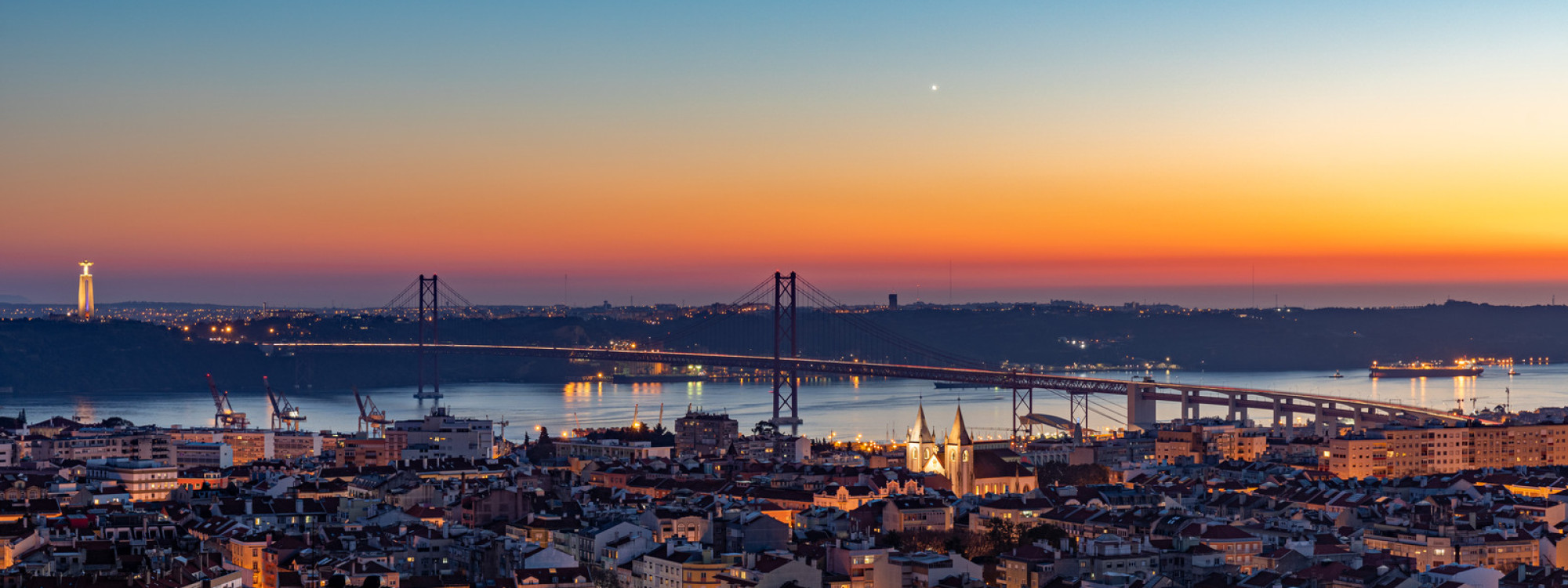Aerial view of the cityscape of Lisbon with illuminated buildings, bridge, and sea during the sunset Aerial view of the cityscape of Lisbon with illuminated buildings, bridge, and sea during the sunset