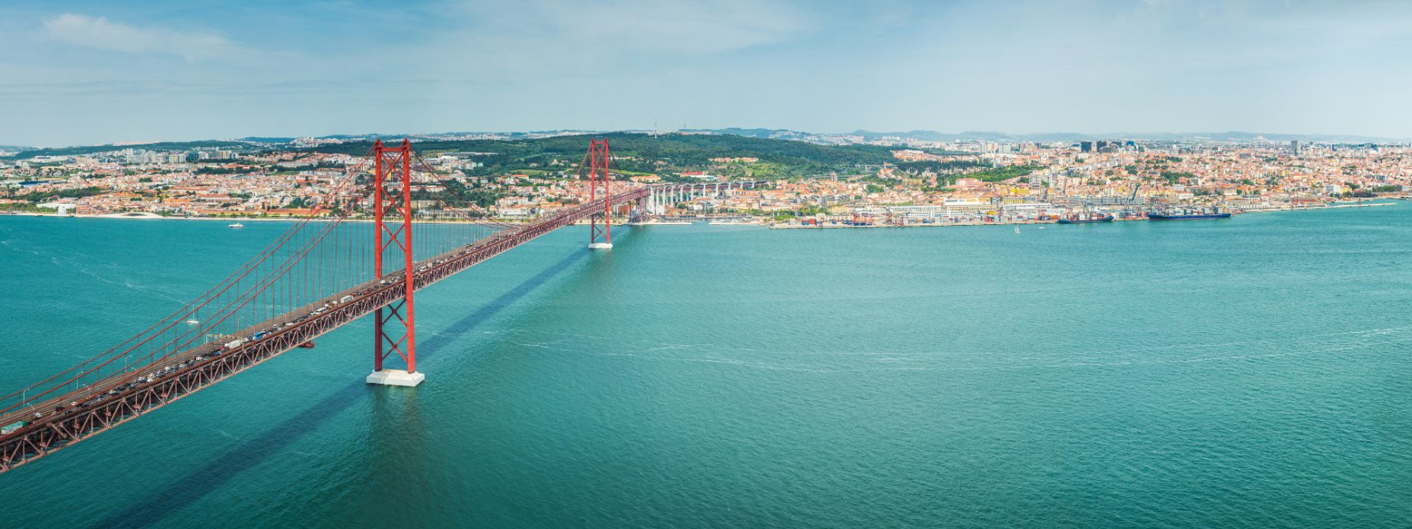 View of the April 25 Bridge in Lisbon, Portugal, with its iconic red color spanning the Tagus River against a blue sky