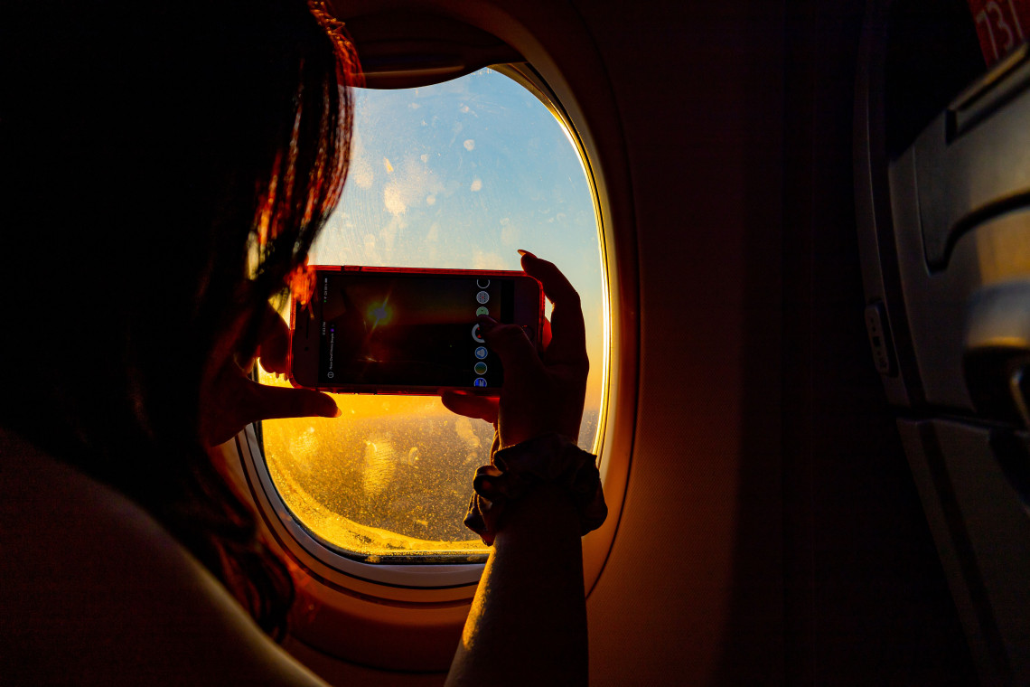 Woman taking a photo of the sunset through an airplane window with her phone during flight