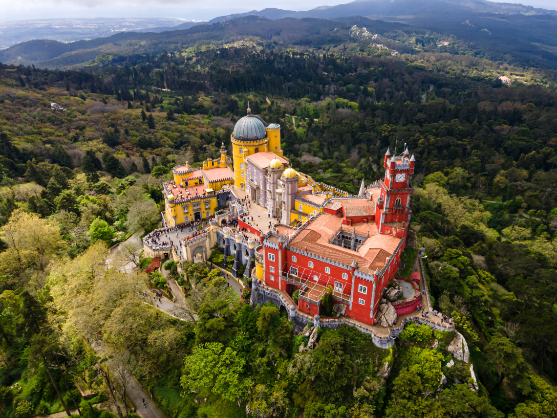 Aerial view of Pena Palace in Sintra, Portugal, with its vibrant red and yellow towers surrounded by lush forested hills.