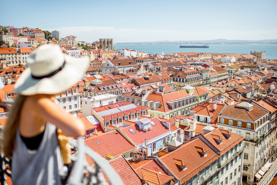 Woman in a sunhat admiring the panoramic view over Lisbon’s red-tiled rooftops and the Tagus River from a scenic viewpoint on a sunny day.