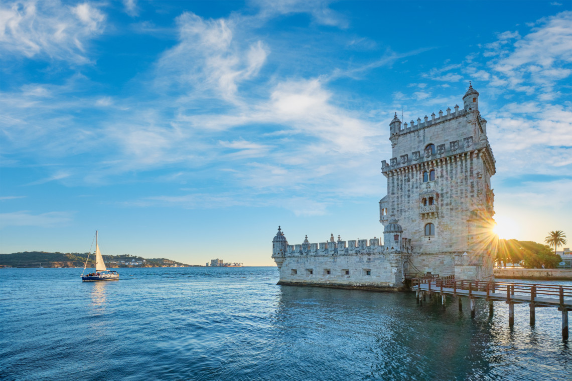 Belem Tower in Lisbon, Portugal, at sunset with a sailboat on the Tagus River and clear blue skies above.