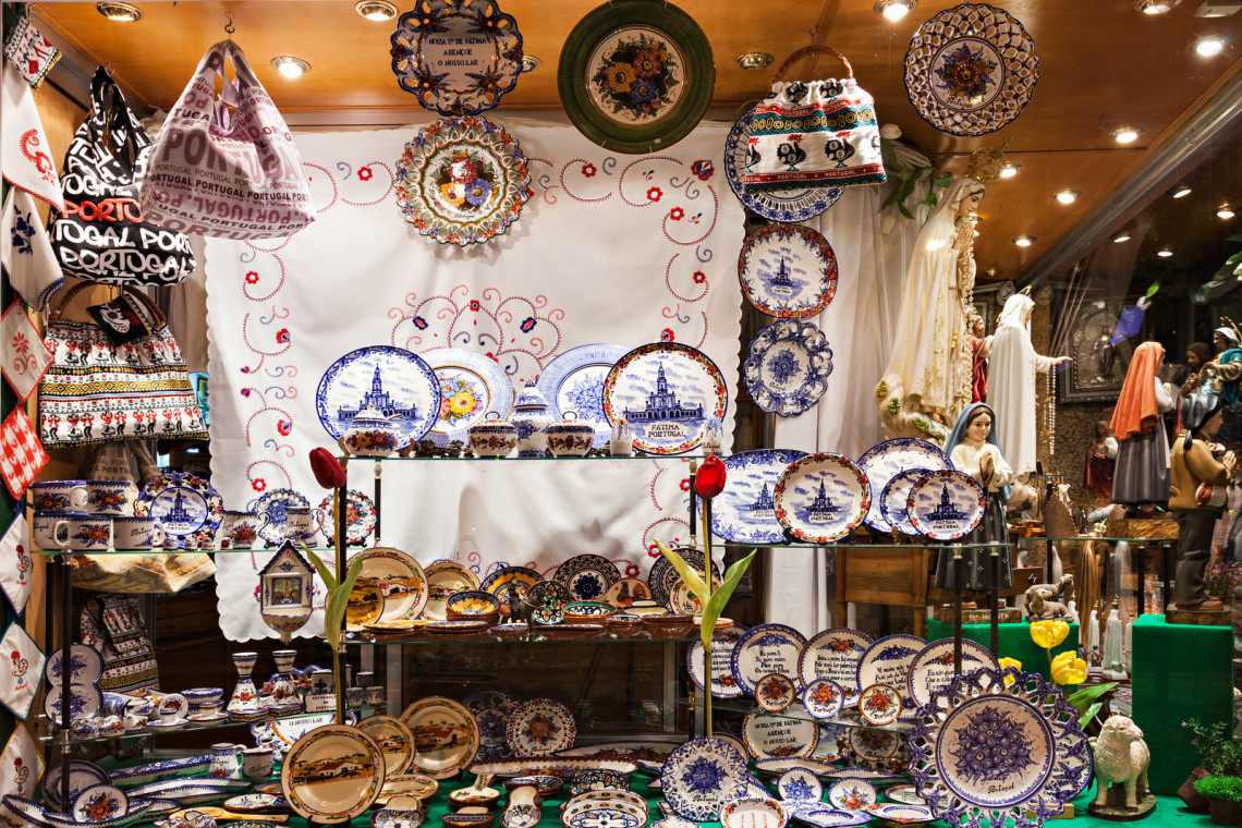 Display of traditional Portuguese souvenirs in Lisbon, including hand-painted ceramic plates, tiles, embroidered textiles, and religious statues, often found in shops near Fátima and central Lisbon.