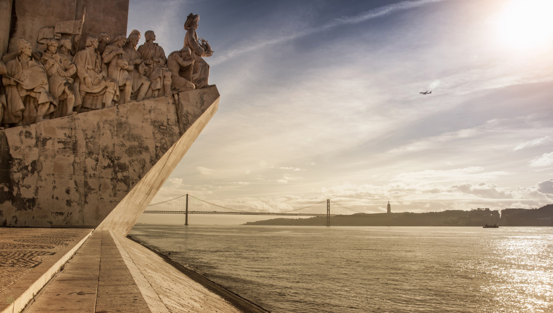 Padrão dos Descobrimentos monument on the waterfront in Belém, Lisbon, with the 25 de Abril Bridge and Cristo Rei statue in the background at sunset.