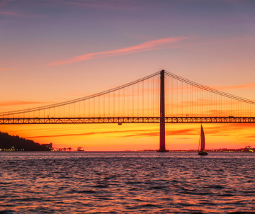 Sailboat on the Tagus River at sunset with the 25 de Abril Bridge in Lisbon, Portugal, silhouetted against a vibrant orange and pink sky.