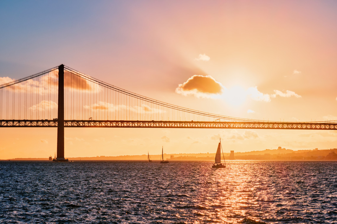 25 de Abril Bridge in Lisbon at sunset with sailboats on the Tagus River, showcasing why Lisbon offers more authentic charm than other Black Friday destinations.