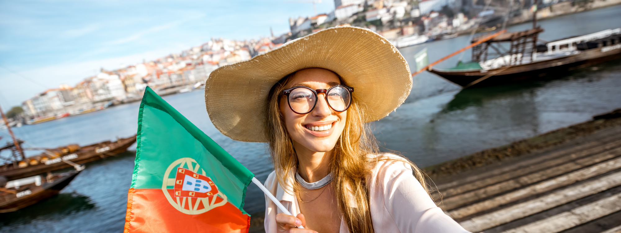 Smiling Traveler with Portuguese Flag in Porto Young woman smiling with a Portuguese flag by the Douro River in Porto, with rabelo boats and the historic city in the background.