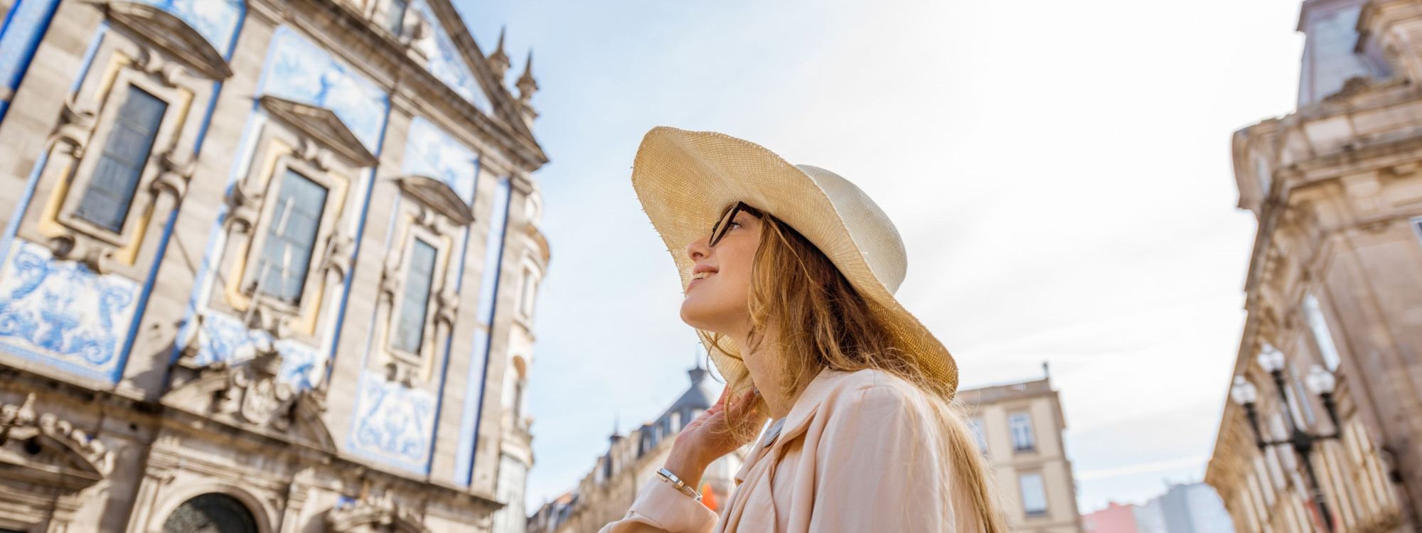 Young Woman Enjoying a Sunny Day in Porto, Portugal – Igreja do Carmo in the Background Woman wearing a wide-brimmed hat and trench coat smiling while exploring the historic center of Porto, Portugal, with the blue-tiled Igreja do Carmo behind her on a sunny day