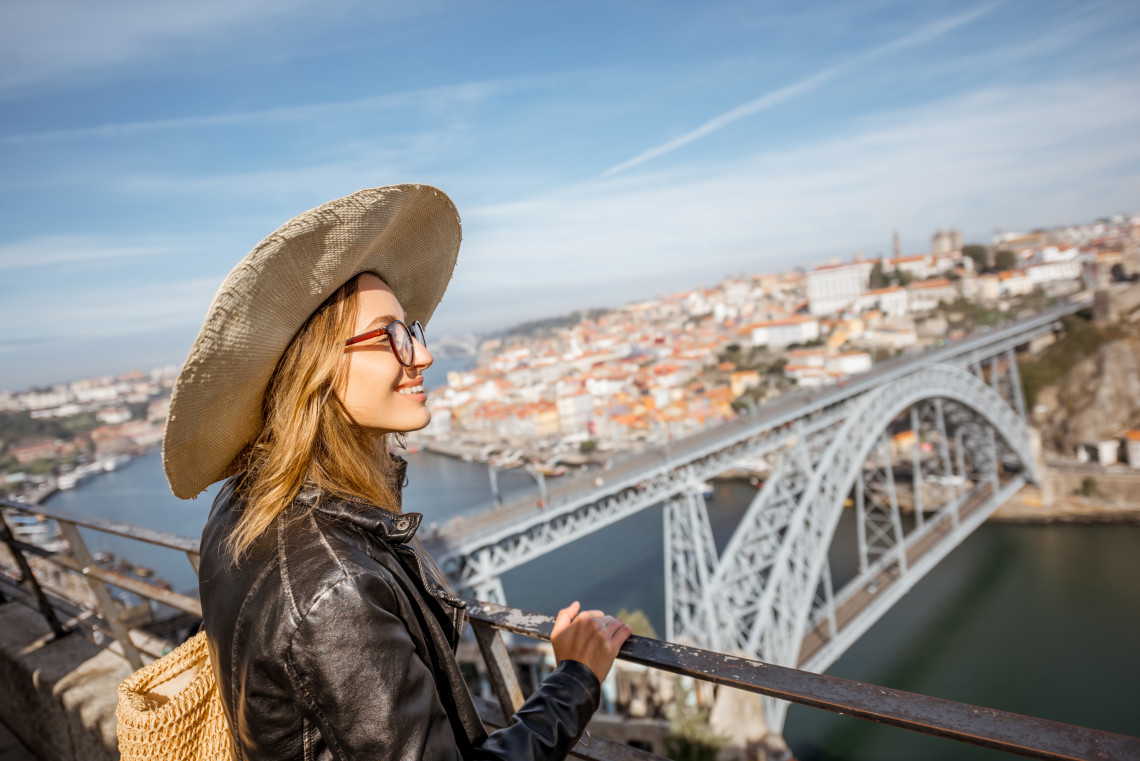Smiling woman wearing a hat and sunglasses enjoying the panoramic view of Porto and the Dom Luís I Bridge from a high vantage point.