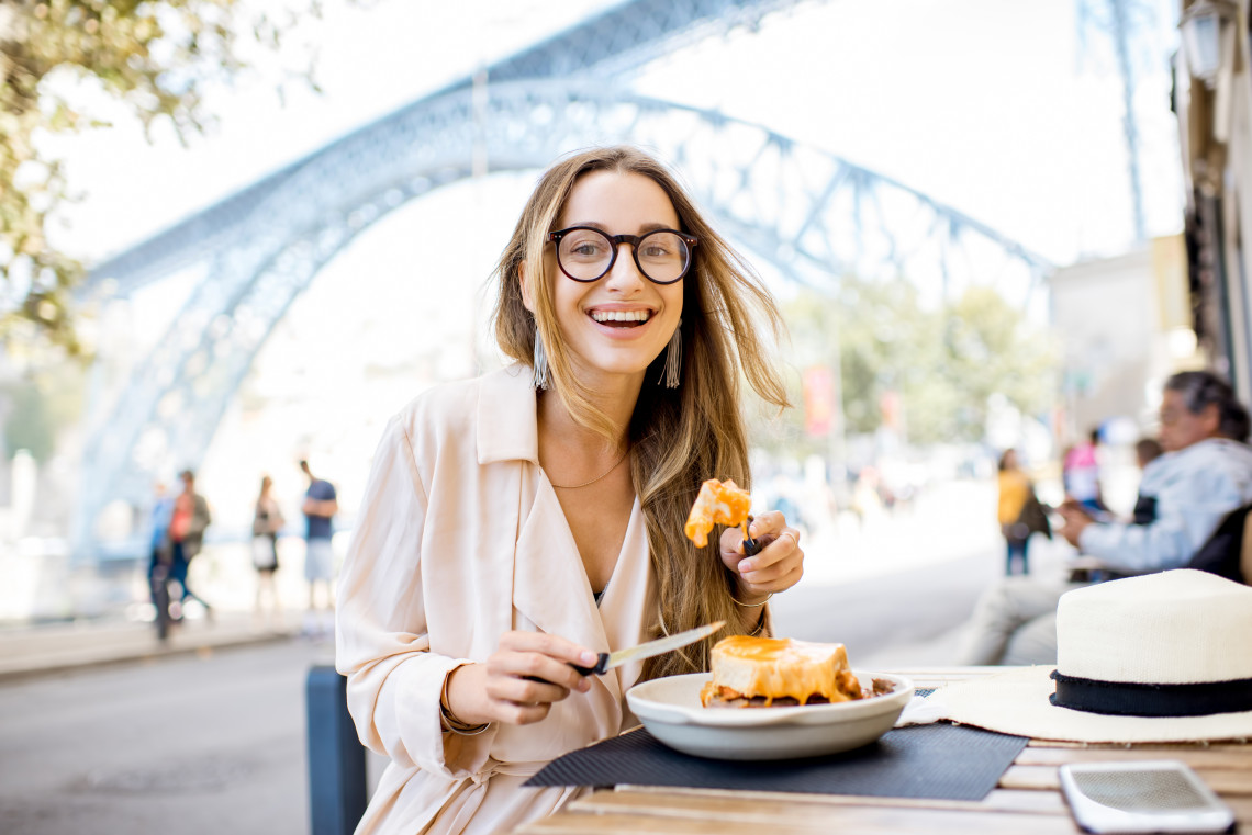 Smiling young woman enjoying a traditional francesinha sandwich at an outdoor café in Porto, with the Dom Luís I Bridge in the background.