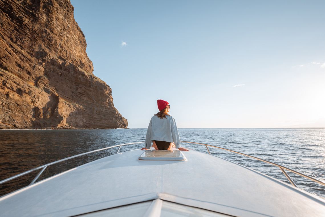Woman sitting at the bow of a boat during a sunset cruise along the dramatic cliffs of the Algarve, Portugal, gazing out at the calm ocean horizon.