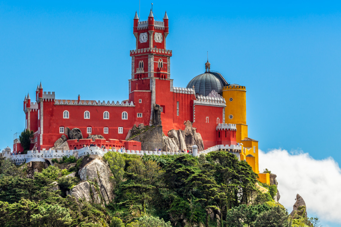 Colorful Pena Palace perched on a hilltop surrounded by lush greenery and clouds in Sintra, Portugal, on a bright sunny day.