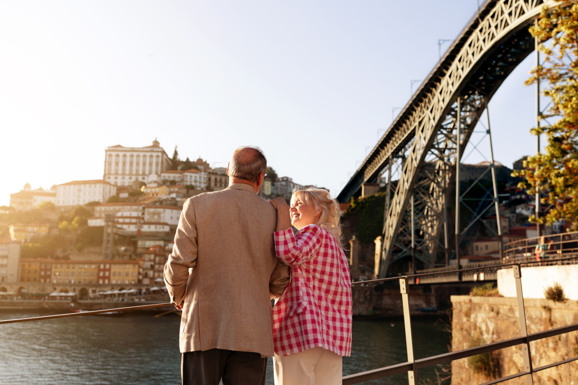 Senior couple enjoying the view of Porto’s historic cityscape and the Dom Luís I Bridge at sunset, standing by the Douro River.