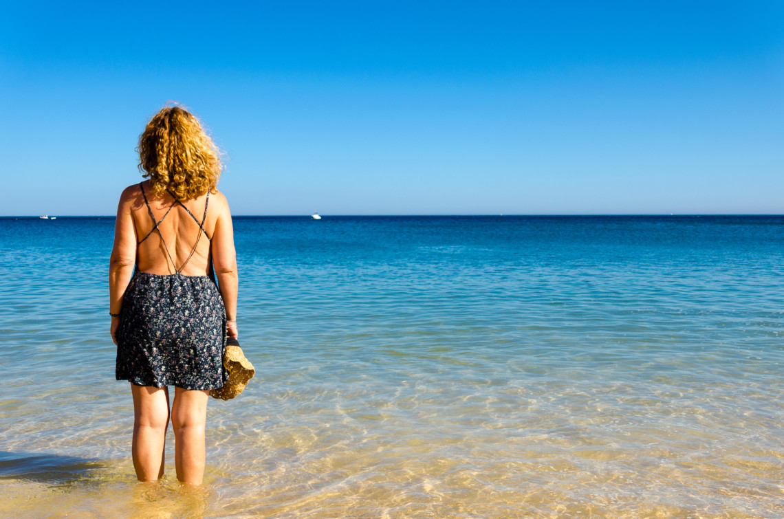 Woman standing at the edge of the clear, calm sea on a sunny day at a beach in the Algarve, Portugal, holding a straw hat and facing the horizon.