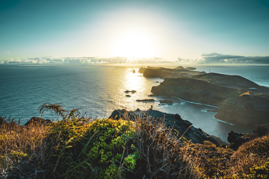 Sitting woman enjoys panoramic view from view point on a steep cliff over the seascape and along the rugged foothills of Madeira coast at sunrise. Ponta do Bode, Madeira Island, Portugal, Europe.