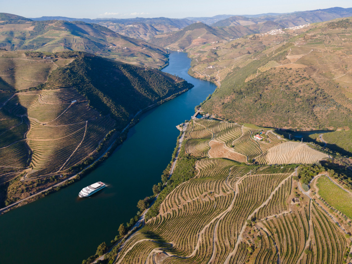 Aerial view of a cruise boat sailing along the Douro River, surrounded by terraced vineyards on rolling hills in Portugal's Douro Valley.