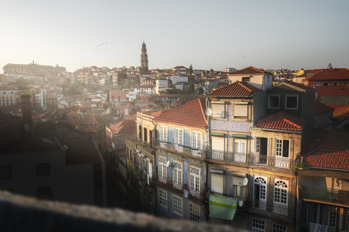 Sunrise over the traditional tiled houses of Porto, Portugal, with Clérigos Tower visible in the background and golden light illuminating the historic cityscape.