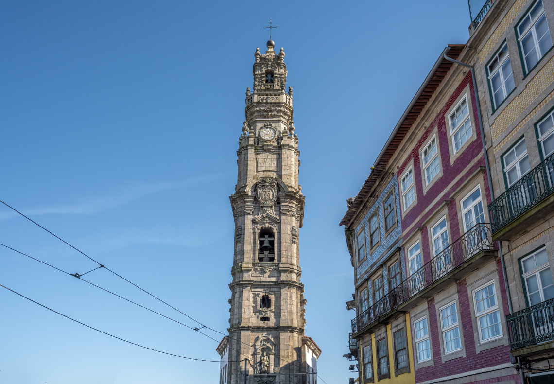 Baroque-style Clérigos Tower in Porto, Portugal, standing tall against a clear blue sky beside colorful traditional Portuguese buildings.