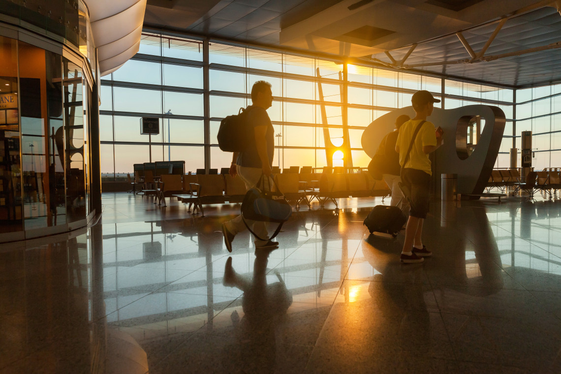 Family walking through a modern airport terminal at sunrise, silhouetted against large glass windows with travel bags.