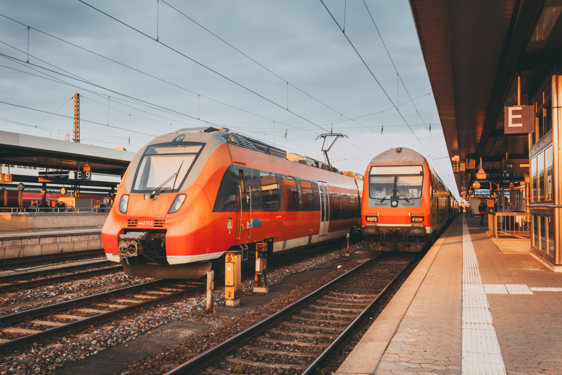 Modern red regional trains stopped at a European railway station platform during golden hour, with overhead electric lines and signage visible.