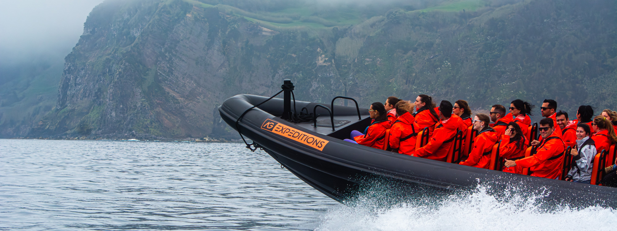AG Expeditions Guests Enjoying a High-Speed Boat Tour Along São Miguel’s Coast Excited group of AG Expeditions passengers in orange jackets raising their hands and smiling during a high-speed boat ride along the cliffs of São Miguel Island’s coastline, Azores