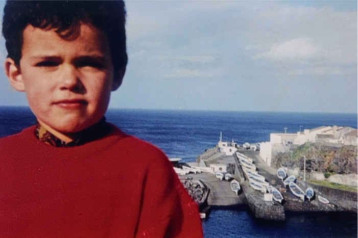 Vintage photo of Luís Nunes as a young boy standing in front of the traditional fishing harbor of Rabo de Peixe on São Miguel Island, Azores, with small boats lined up on the pier and the Atlantic Ocean in the background