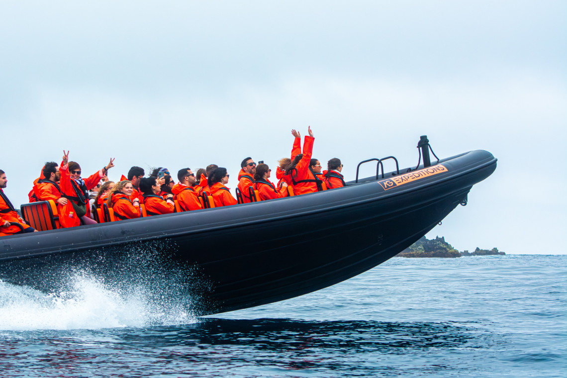 Passengers in orange jackets enjoying a thrilling AG Expeditions speed boat ride off the coast of São Miguel Island, Azores, with water splashing as the boat lifts above the waves and rocky islets in the distance