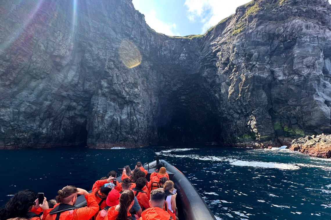 Passengers on an AG Expeditions boat approaching a dramatic sea cave along the volcanic cliffs of São Miguel Island, Azores.