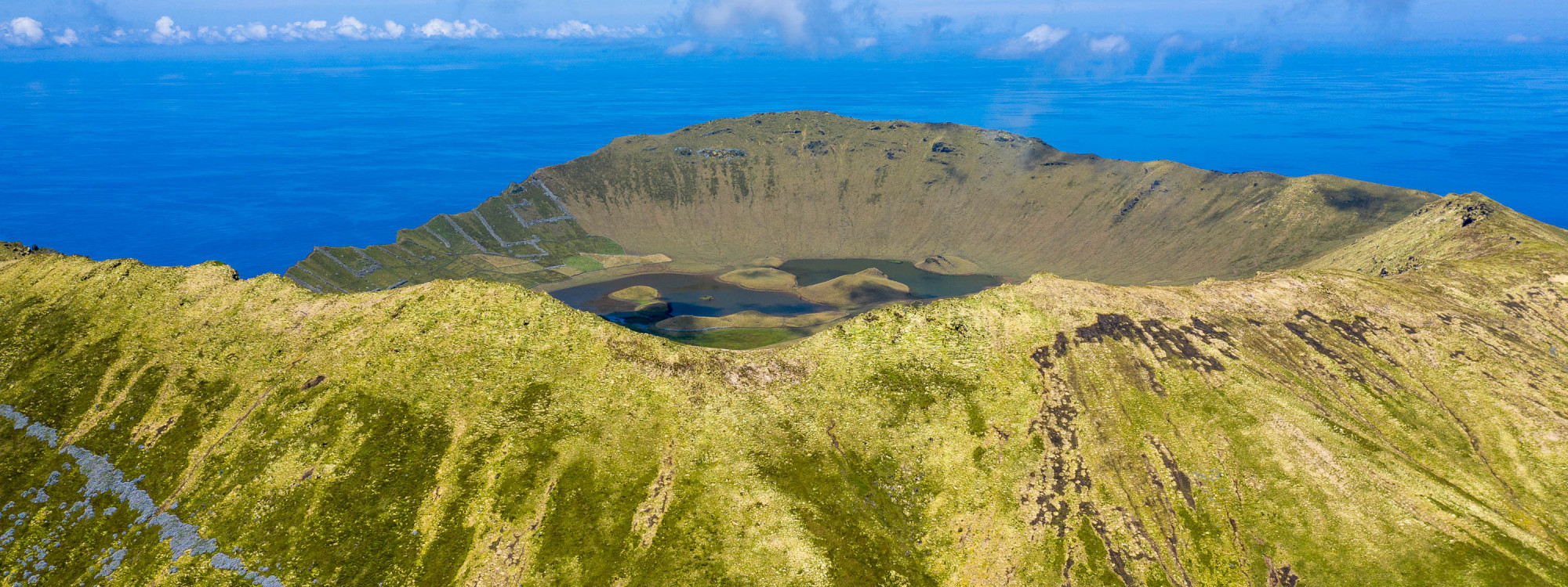 Panoramic view of Caldeirão Crater on Corvo Island in the Azores, Portugal. Small lakes and volcanic formations occupy the caldera floor, surrounded by green crater walls and the Atlantic Ocean.