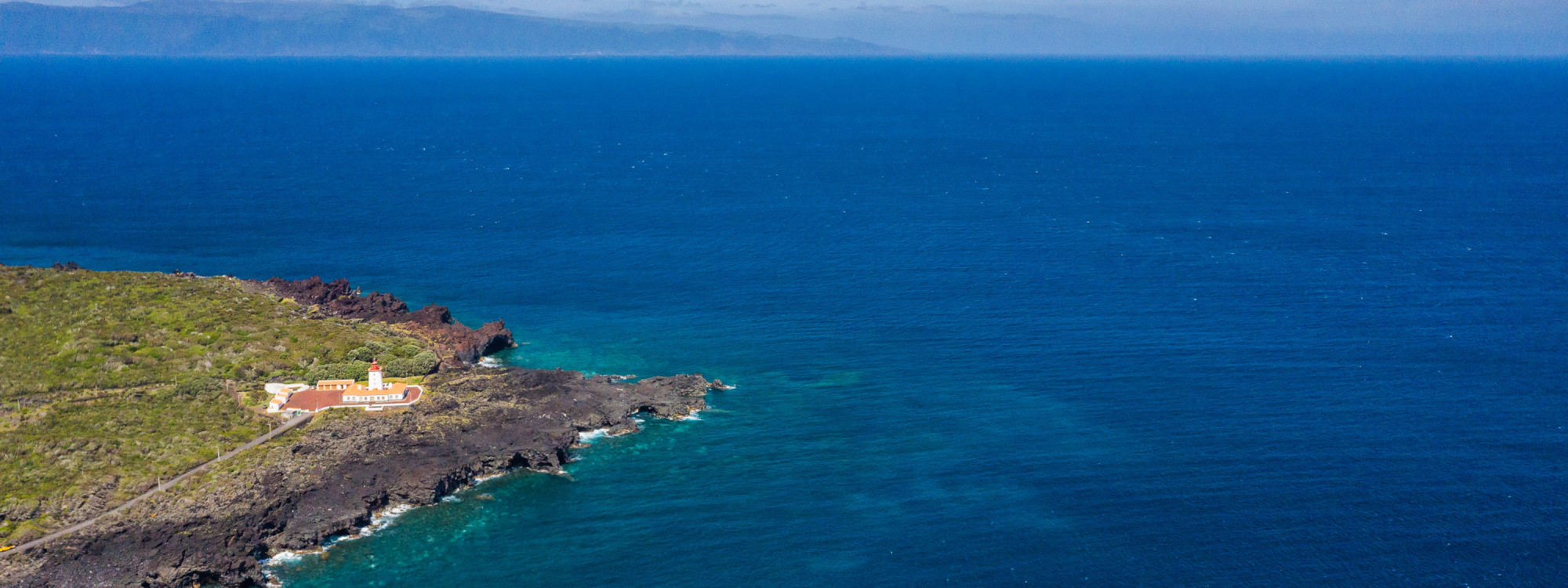 Aerial view of a volcanic headland on Pico Island in the Azores, extending into the deep blue Atlantic Ocean with clear turquoise water along the rocky shoreline.
