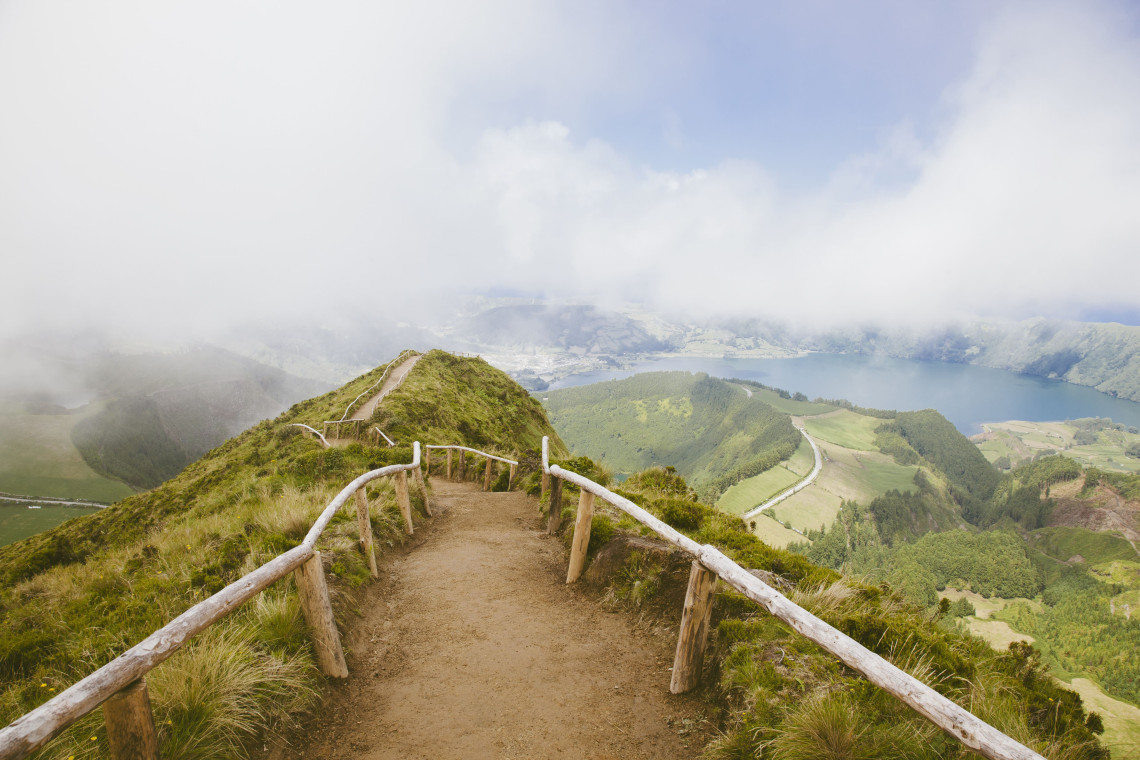 Dirt trail with wooden railings leading to Boca do Inferno viewpoint, overlooking the lush green crater of Sete Cidades on São Miguel Island, Azores, partially covered by mist.