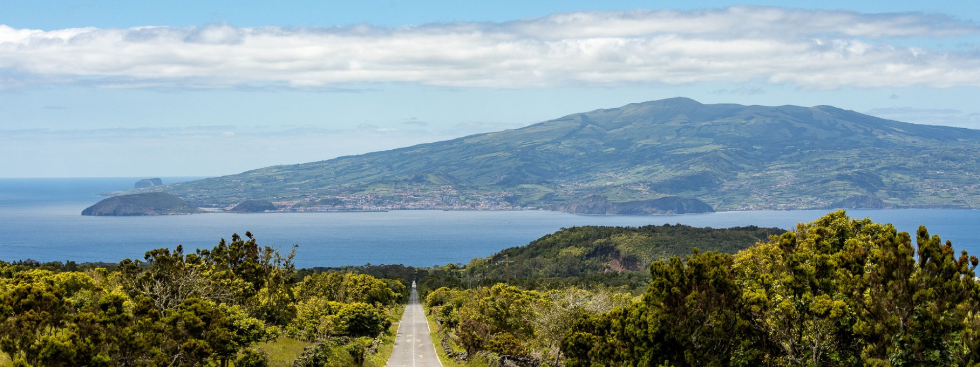 A straight road cutting through dense green vegetation on Faial Island, leading toward the Atlantic channel with the volcanic mass of Pico Island and its peak rising across the water under a partly cloudy sky