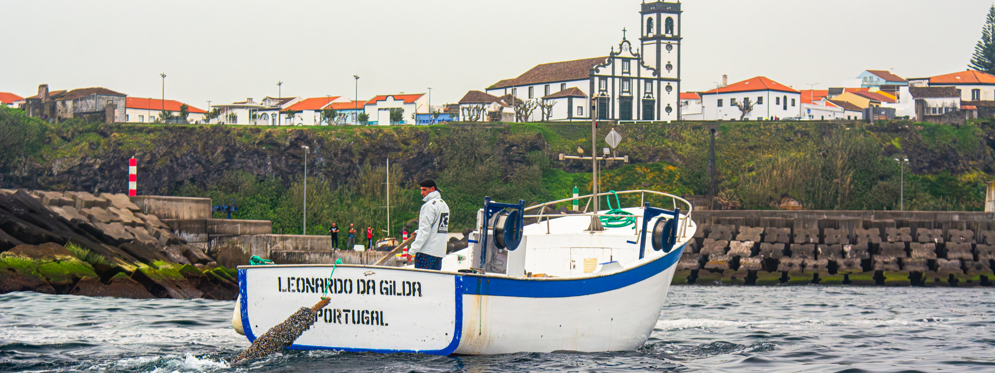 Barco de Pesca em Rabo de Peixe – Tradição Marítima nos Açores Barco de pesca tradicional no porto de Rabo de Peixe, com vista para a vila e a igreja ao fundo, na ilha de São Miguel, Açores.