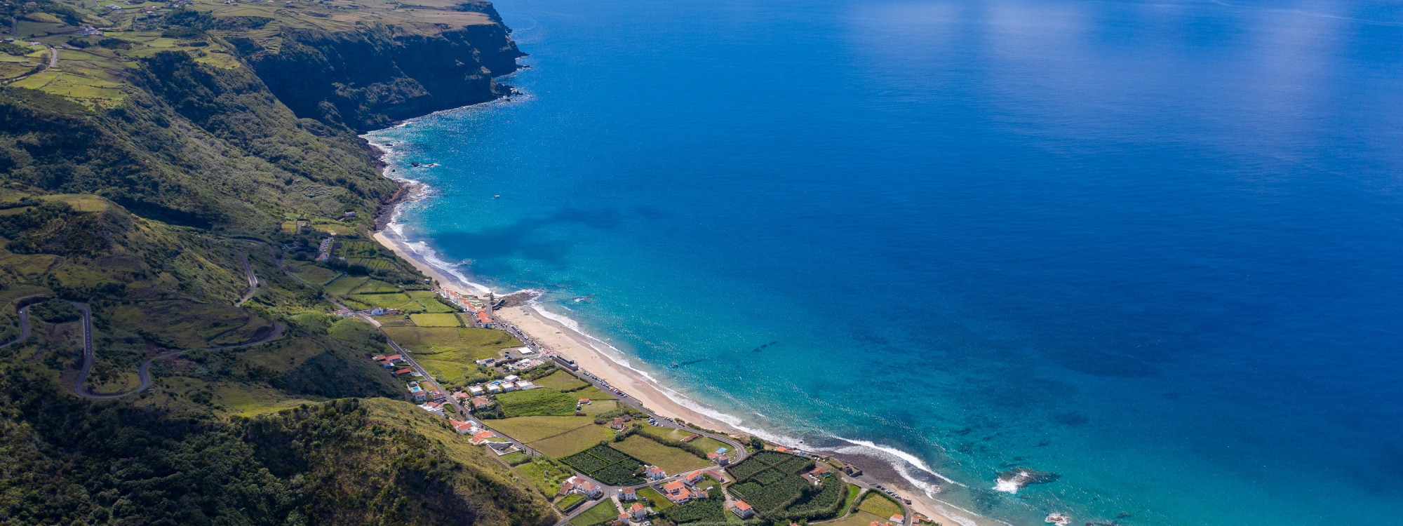 Aerial view of Praia Formosa beach on Santa Maria Island, Azores, with turquoise Atlantic water, a long sandy beach, and green terraced agricultural fields climbing the coastal hillside above