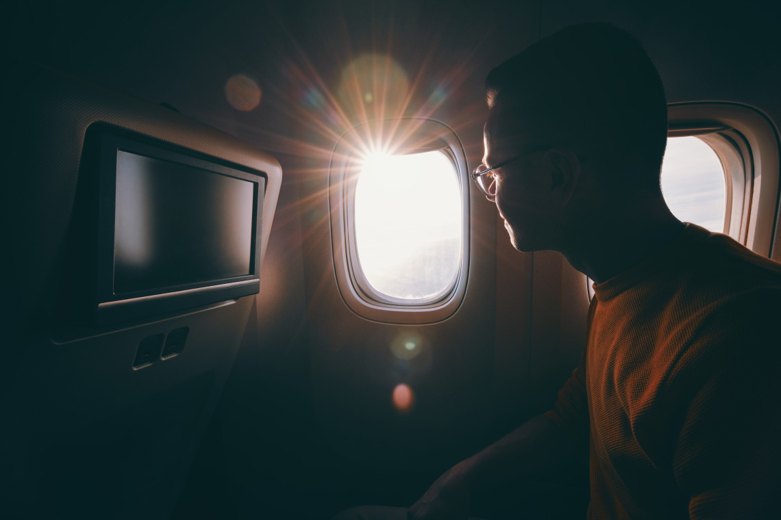 Man gazing out of an airplane window at sunrise, with sun rays entering the cabin and an entertainment screen visible on the seatback.