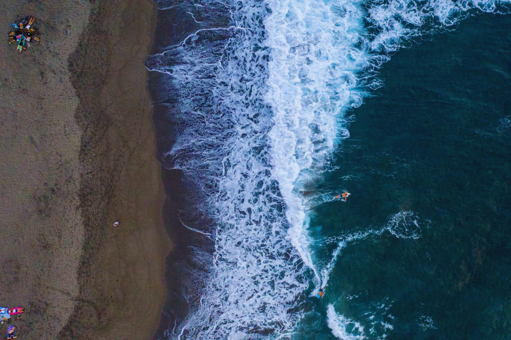 Aerial view of Santa Bárbara Beach in São Miguel Island, Azores, showing dark sand, foamy waves, and surfers in the ocean near the shoreline.