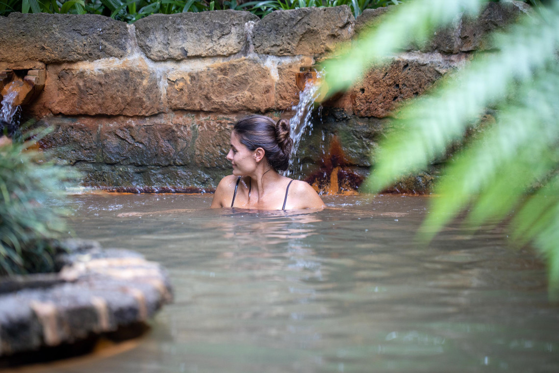 Woman relaxing in a natural hot spring pool at Terra Nostra Park in Furnas, São Miguel Island, surrounded by stone walls and tropical plants.