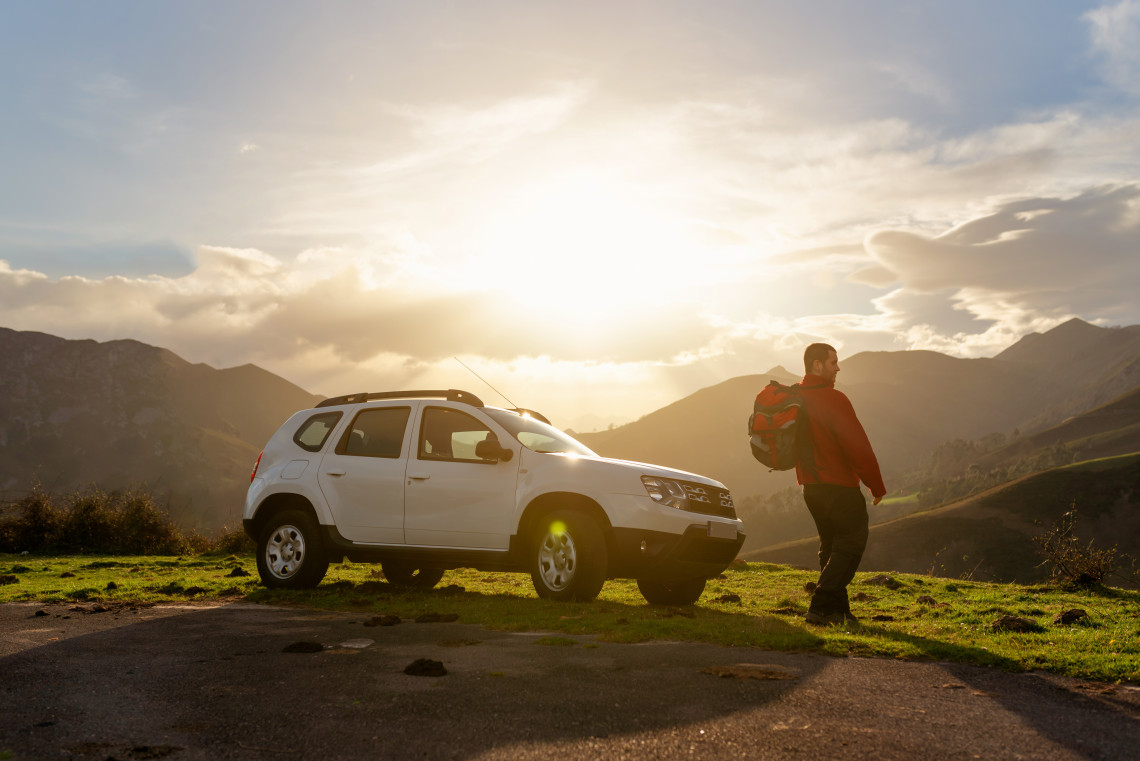 Man with a backpack standing beside a white SUV at sunset, surrounded by mountains and green hills on São Miguel Island, Azores.