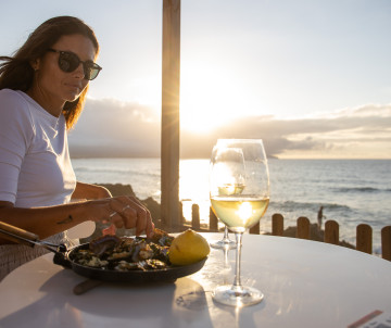 Woman enjoying seafood and white wine at a seaside restaurant near Santa Bárbara Beach in São Miguel Island, Azores, with the sun setting over the Atlantic Ocean.