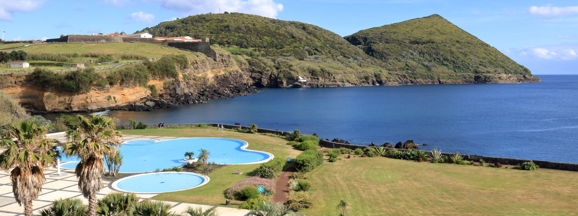 Outdoor swimming pool and landscaped grounds at Hotel do Caracol in Angra do Heroísmo, overlooking the Atlantic Ocean and the green slopes of Monte Brasil.