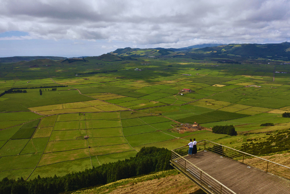 Two friends standing at Serra do Cume viewpoint in Terceira Island, Azores, overlooking a vast green patchwork of farmlands under a cloudy sky.