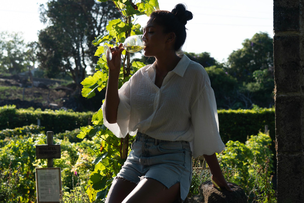 Woman enjoying a glass of white wine at a vineyard in Biscoitos, Terceira Island, Azores, surrounded by lush green vines and volcanic stone walls.