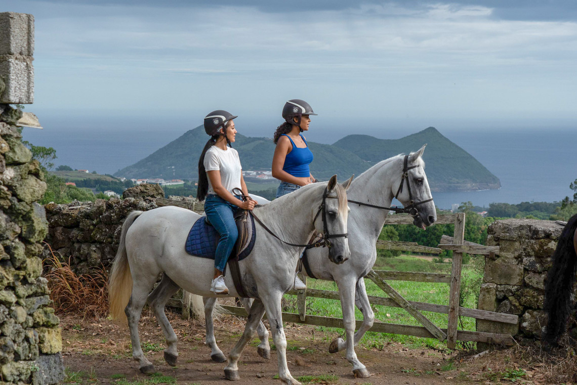 Two women horseback riding near a stone wall on Terceira Island, Azores, with scenic ocean views and Monte Brasil rising in the background